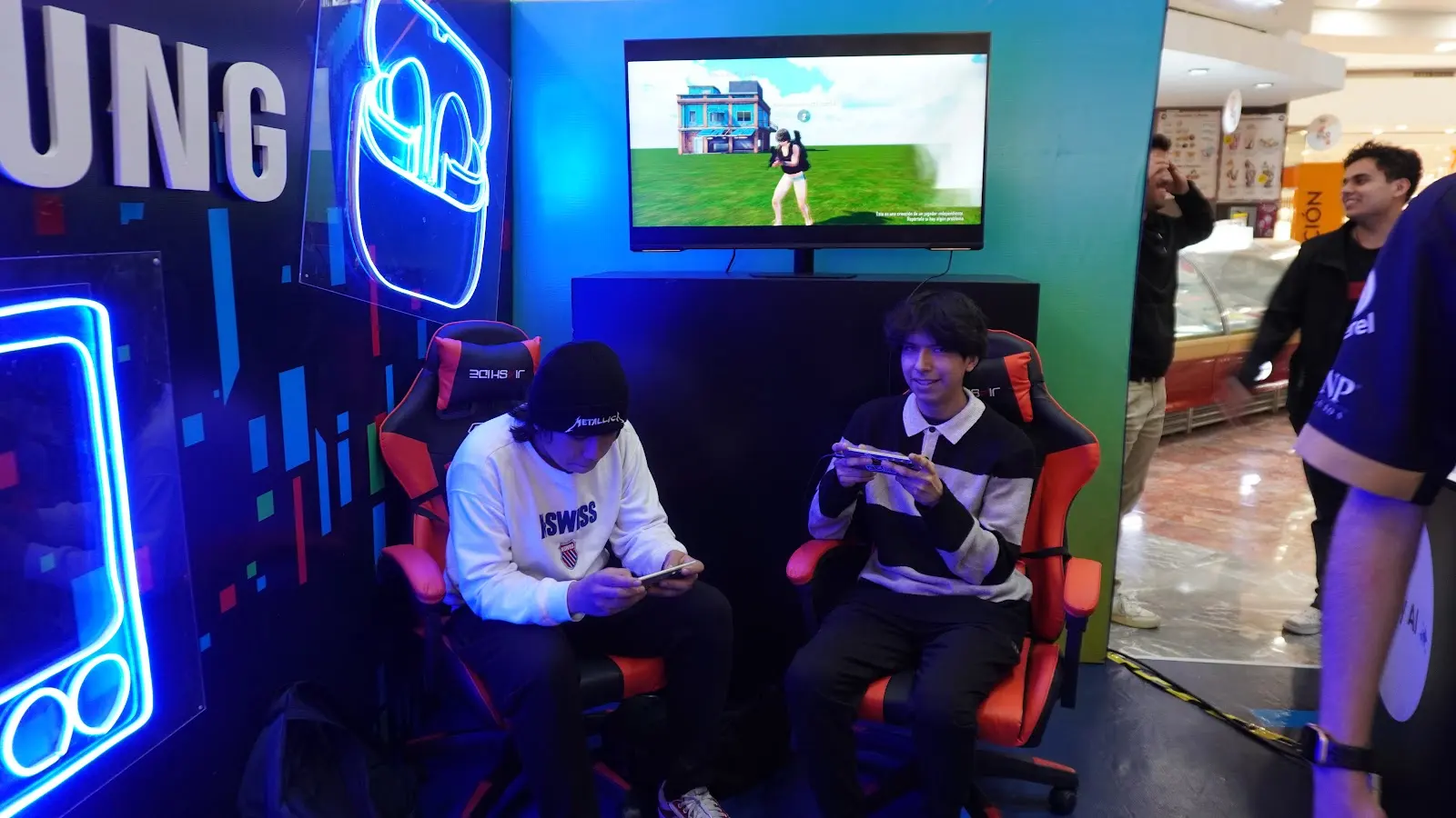 Dos participantes juegan en dispositivos móviles dentro de una activación Samsung, con iluminación azul y pantalla de PUBG MOBILE